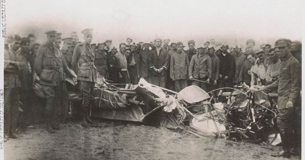 The crumpled remains of a plane's fuselage on sand, surrounded by men in uniform.